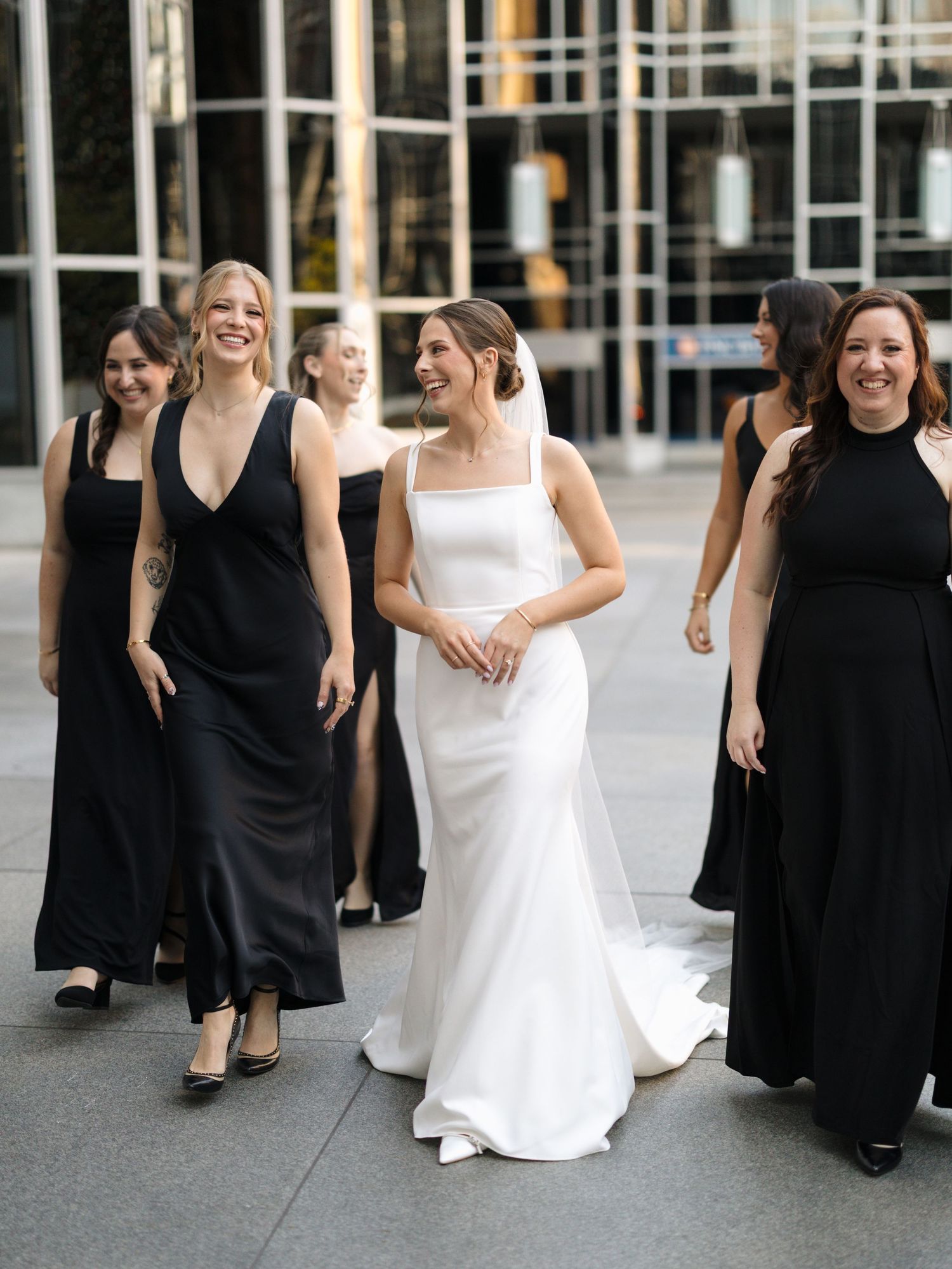 Bridesmaids in black dresses walking with bride in white gown during outdoor wedding.