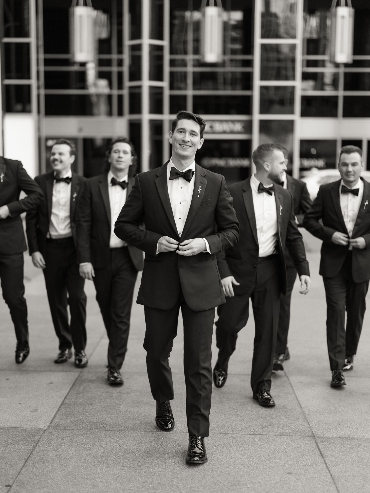 Groomsmen in black tuxedos walk in formation along a modern building exterior.