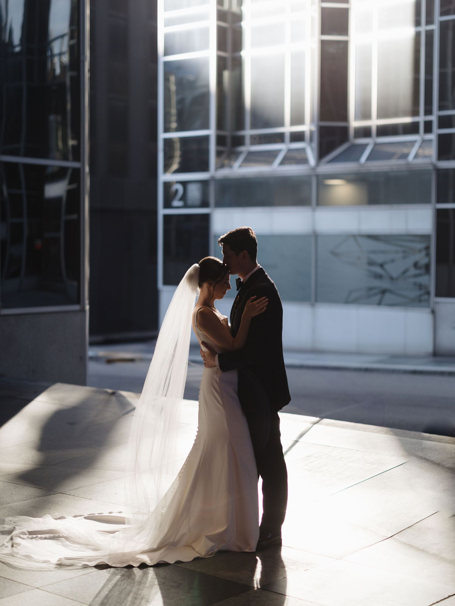 Bride in flowing white gown and groom share tender moment in dramatic window lighting.