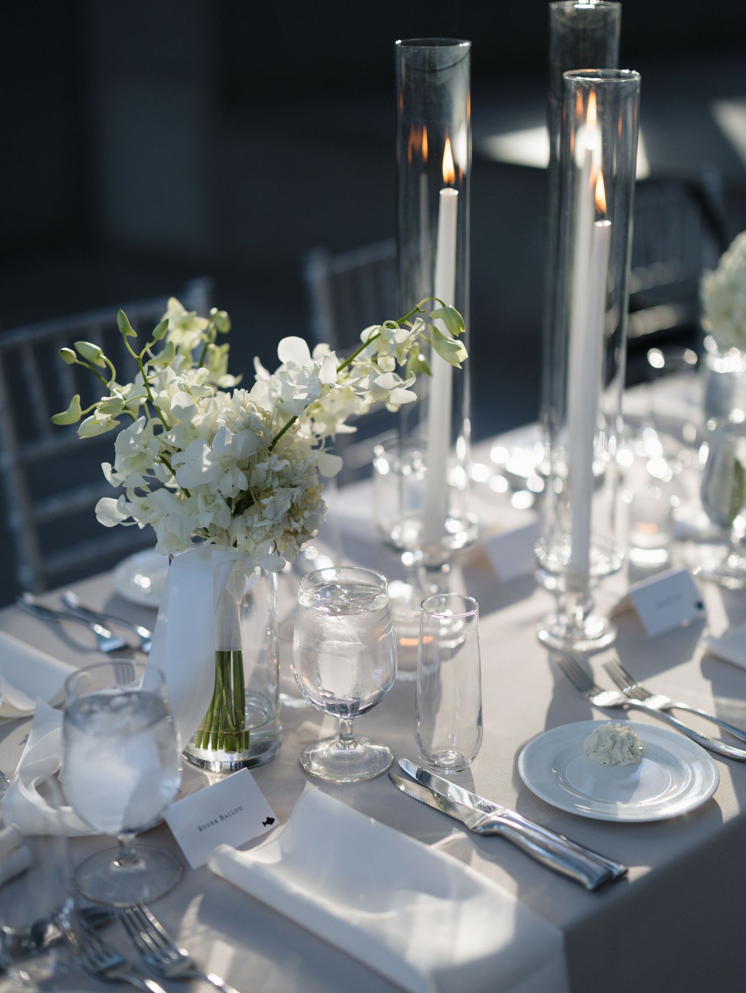Elegant white table settings with crystal glassware, candles and delicate floral arrangements in natural sunlight.