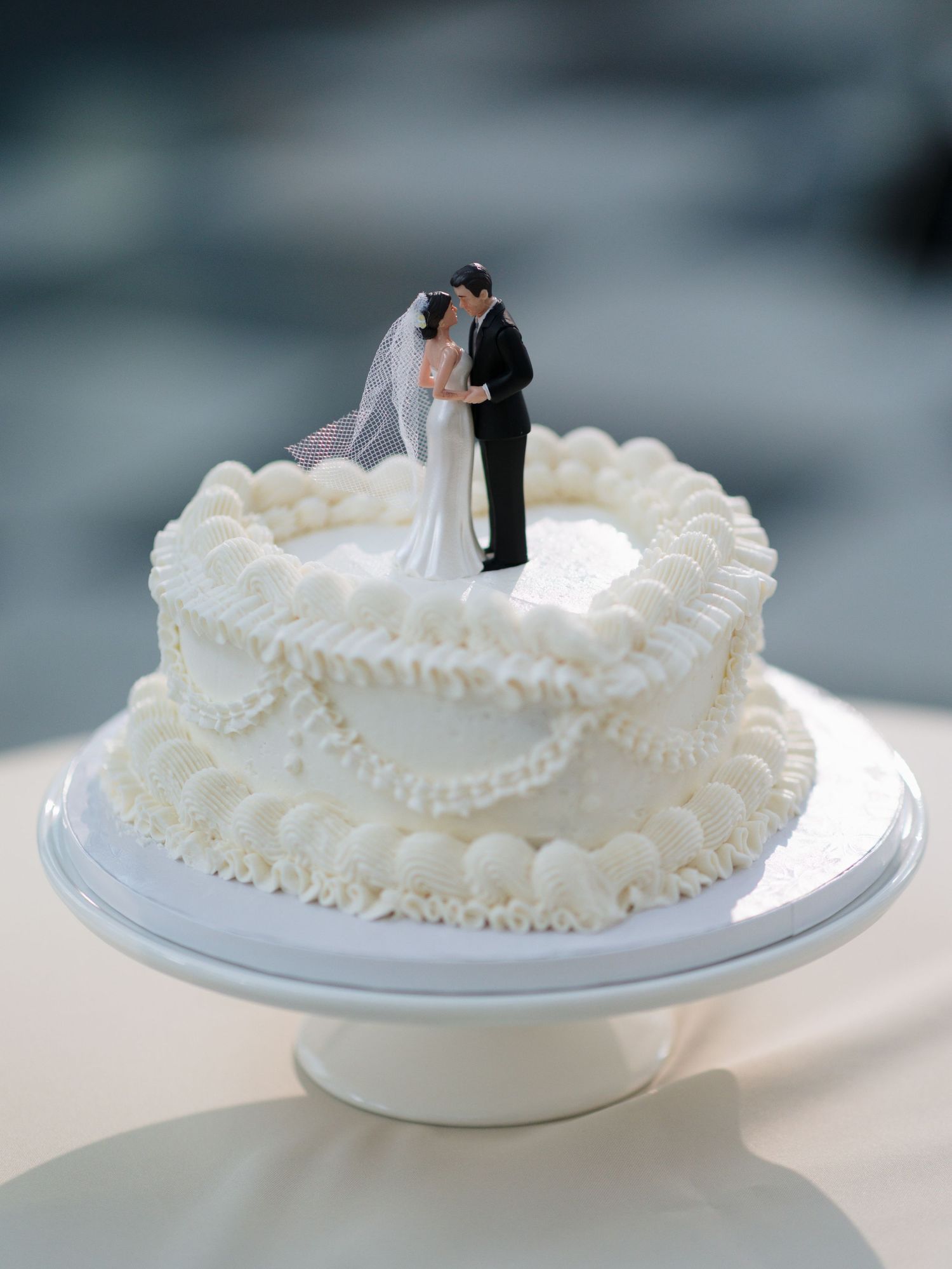 A white two-tiered wedding cake decorated with delicate piping and topped with a traditional bride and groom figurine.