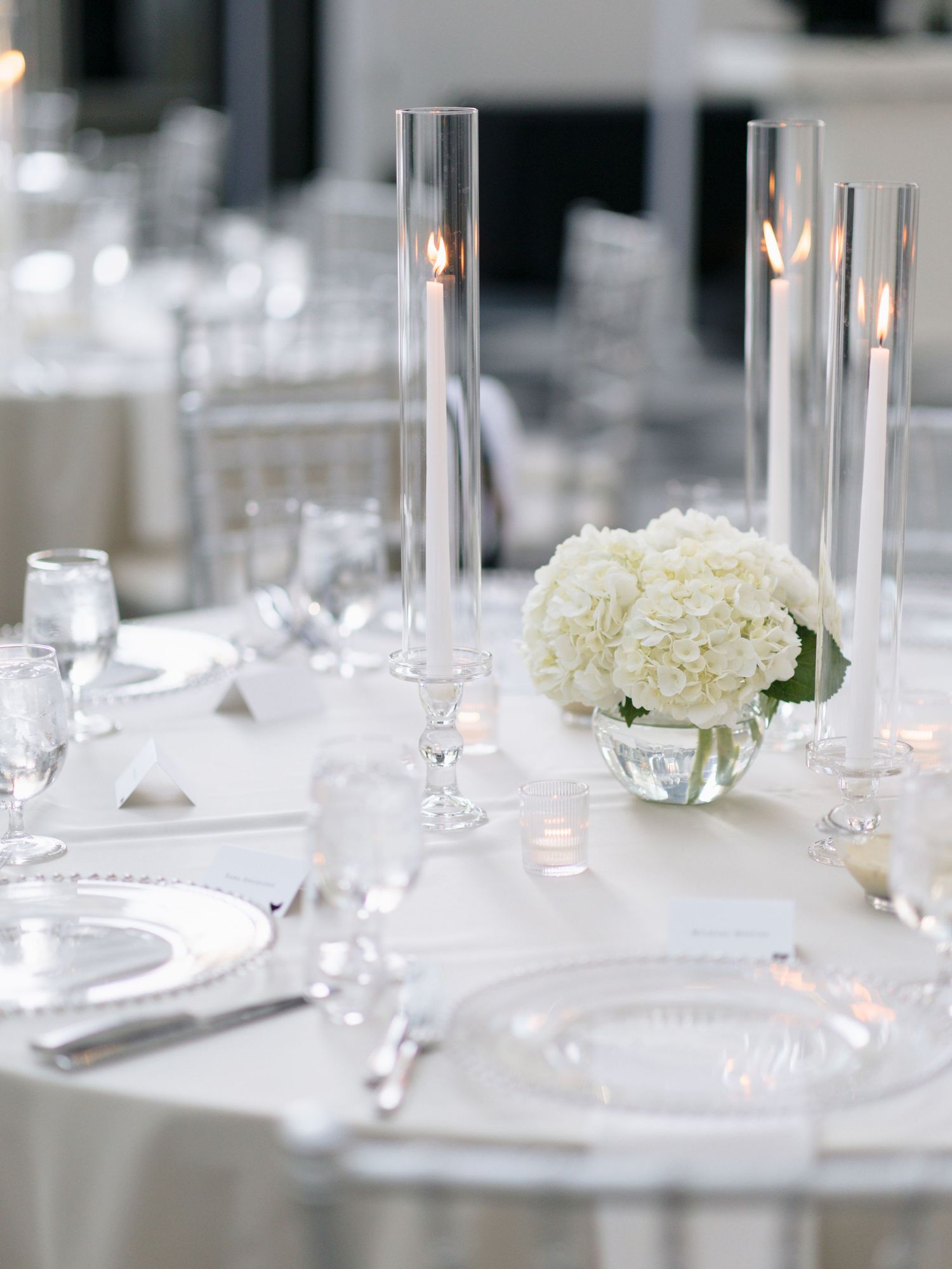 Minimalist white wedding reception table setting with glass vases and white hydrangea centerpieces.