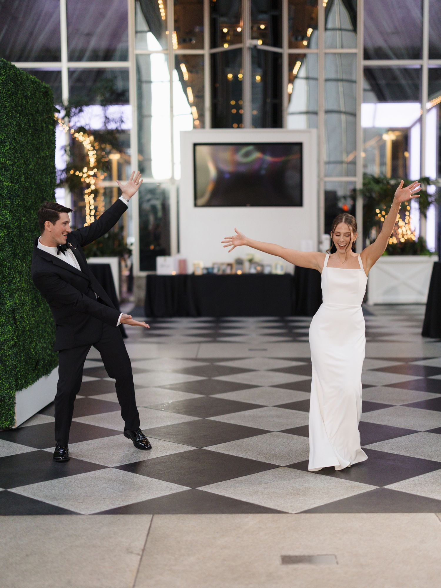 A couple performs a playful dance sequence on a black and white checkered floor with string lights in the background.