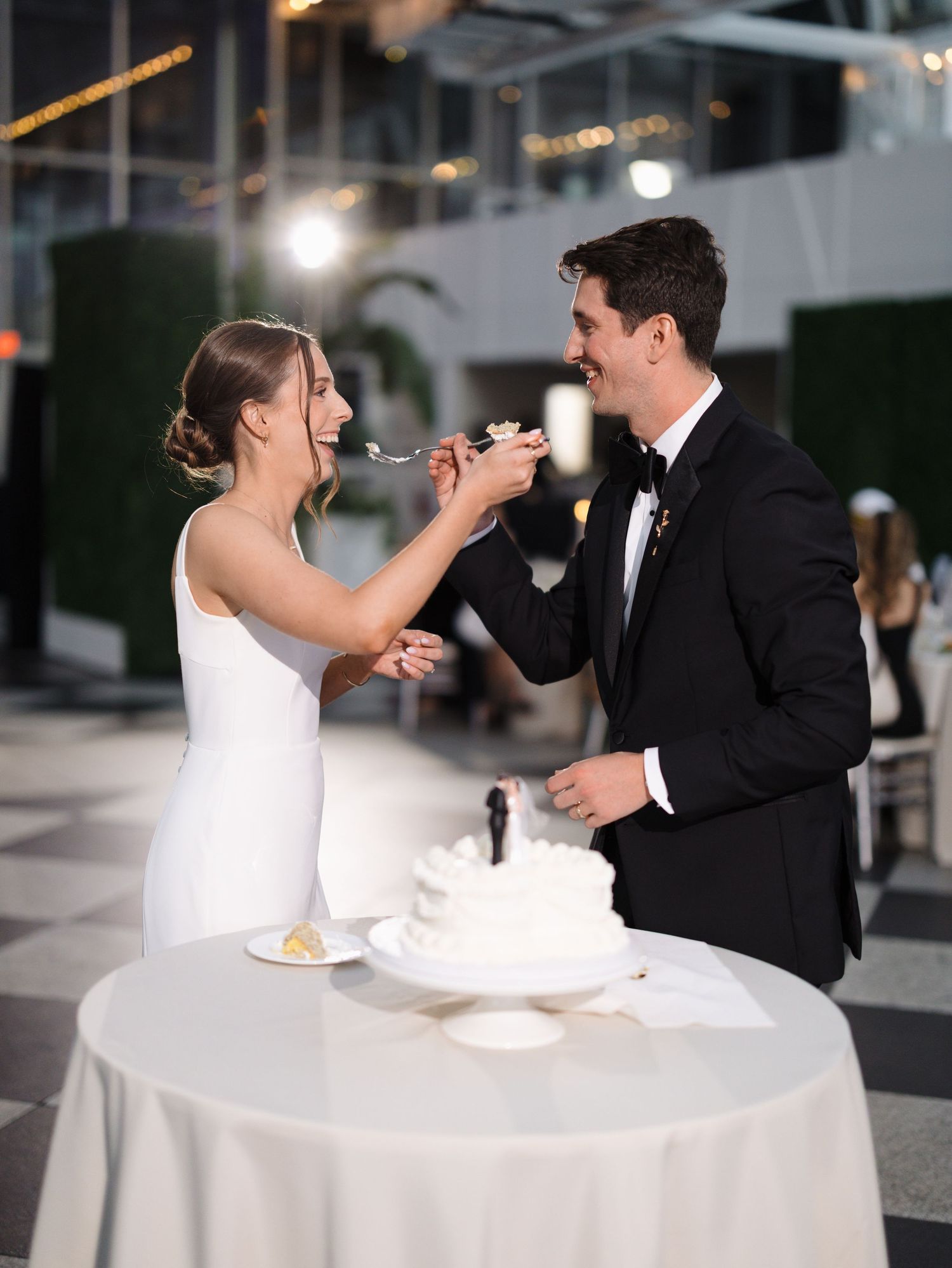 Newlywed couple playfully smashes cake into each other's faces during wedding reception at an elegant venue.