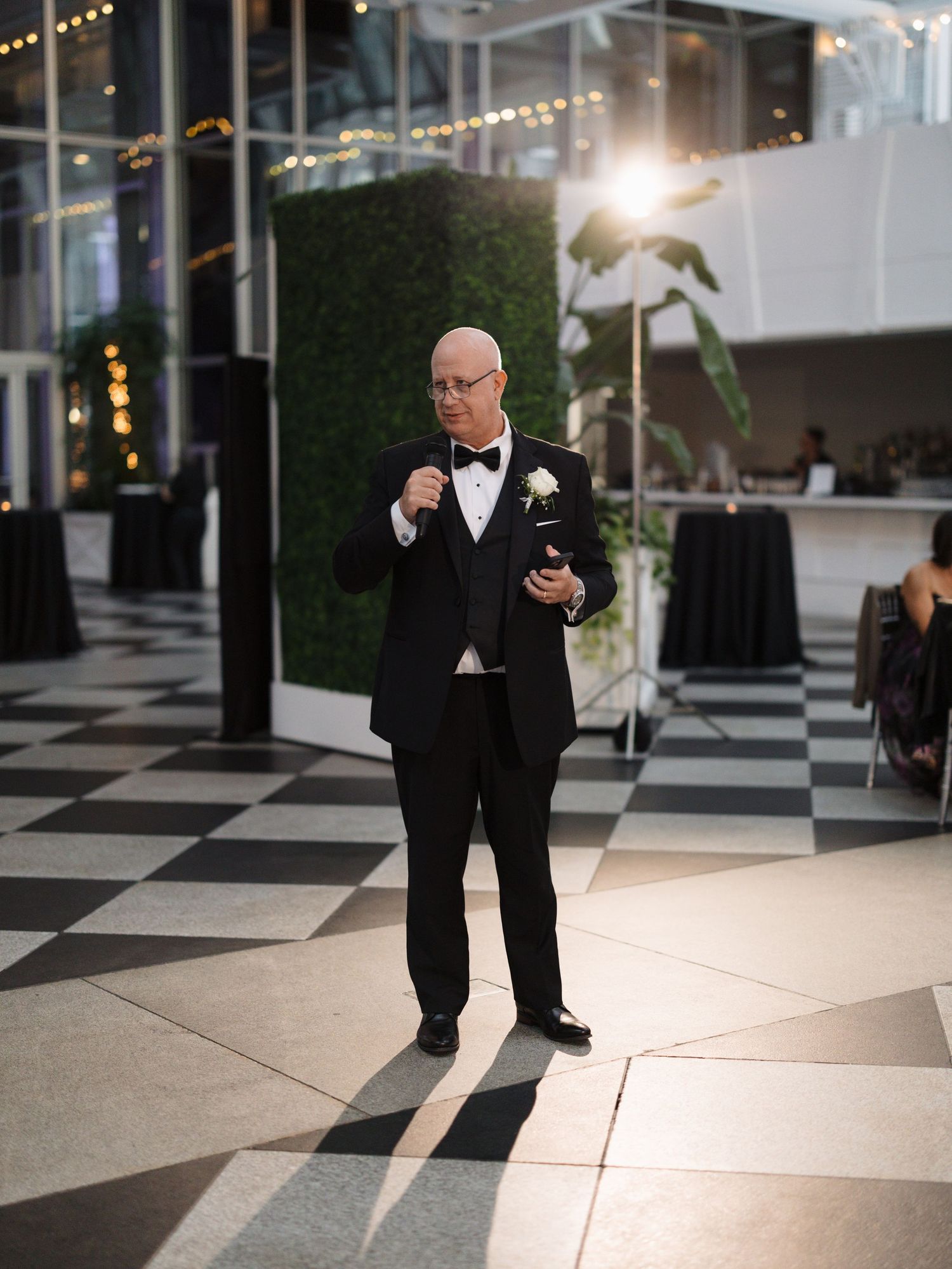 A person in a black suit checks their phone while standing on a checkered floor under string lights.