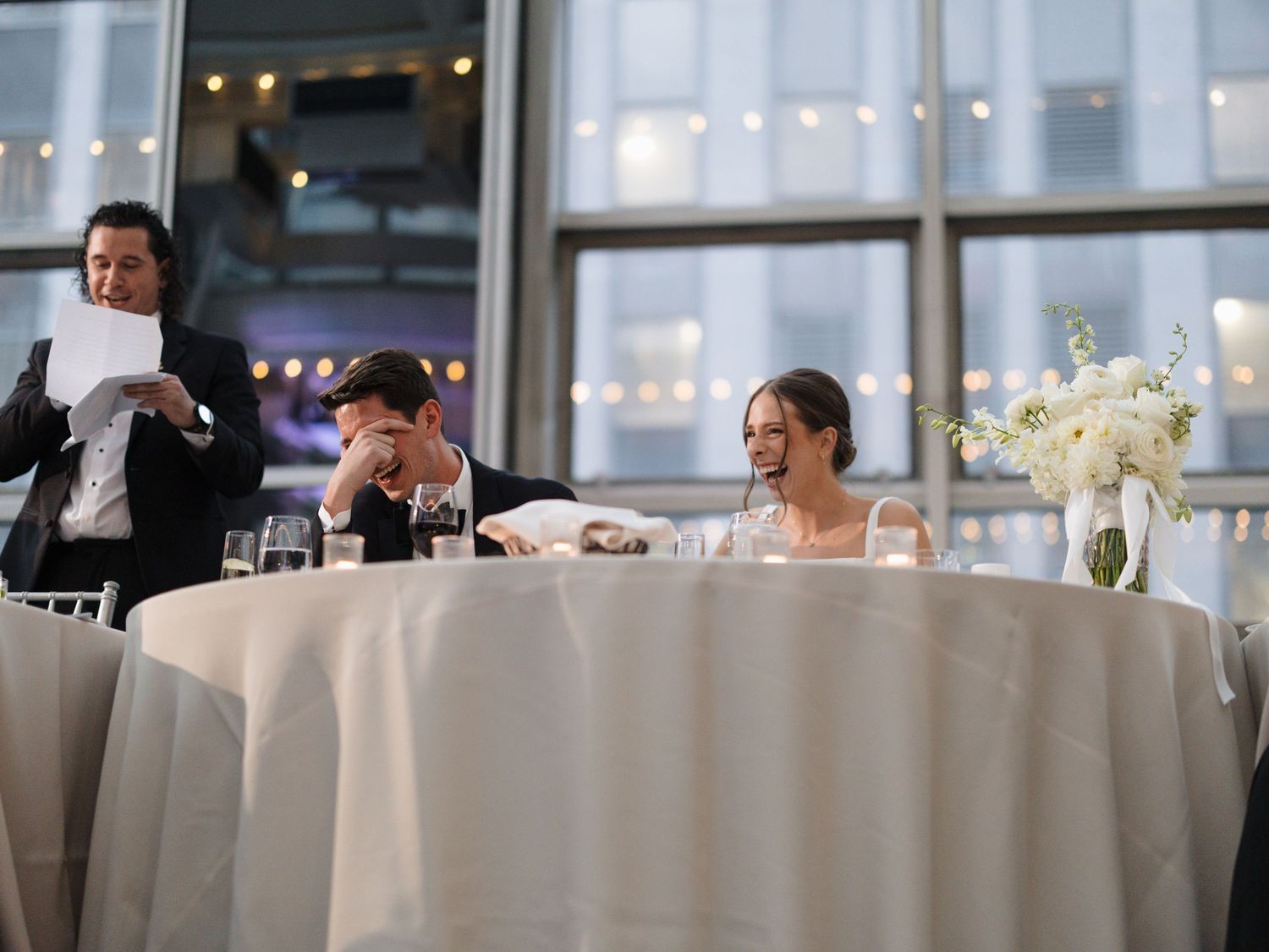 Elegant wedding reception venue with white tablecloths and floral centerpieces in a bright room with large windows.
