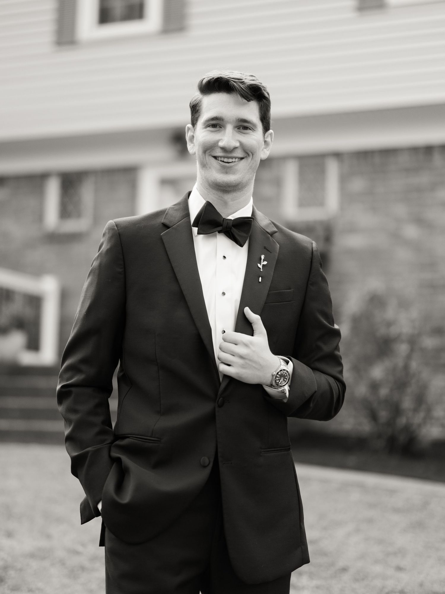 A black and white photo of someone in a tuxedo and bowtie smiling while adjusting their cufflinks.