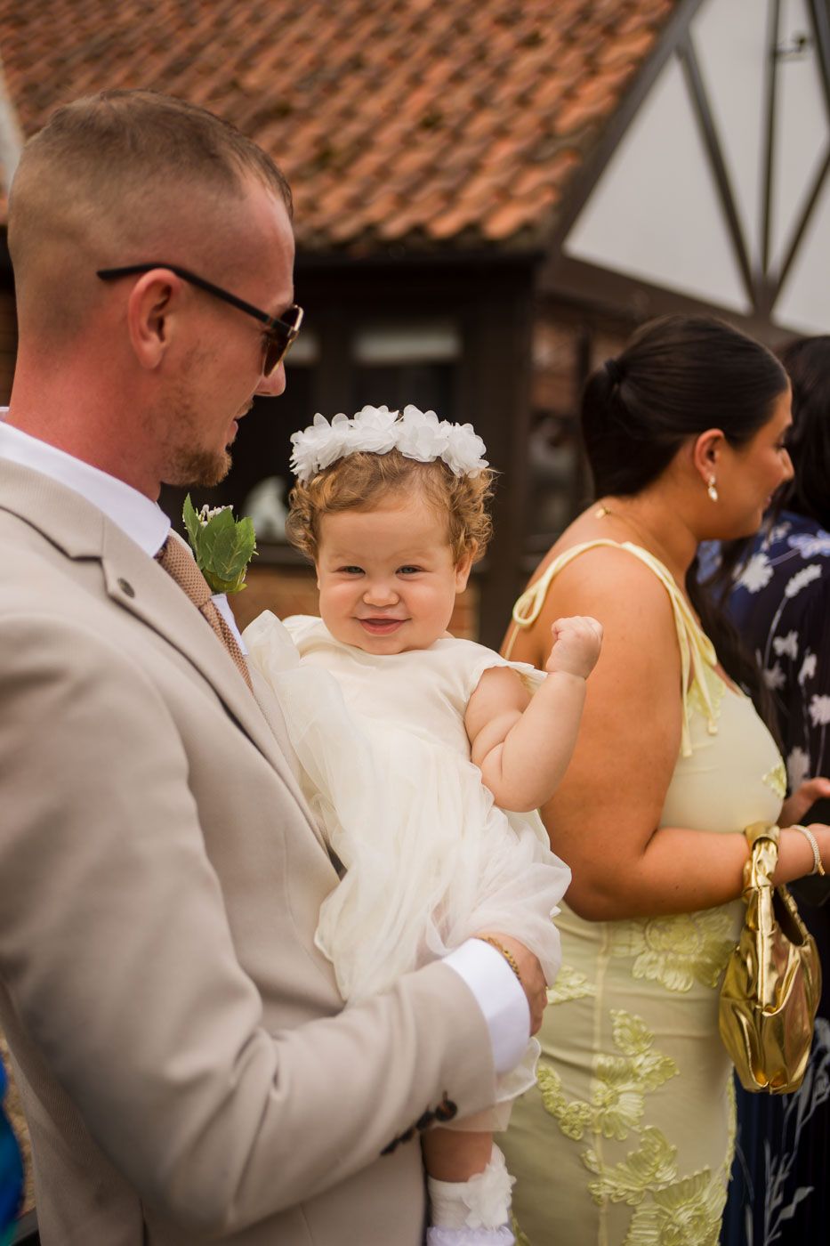 Tender family moment captured at a wedding with a baby in white dress and floral crown.