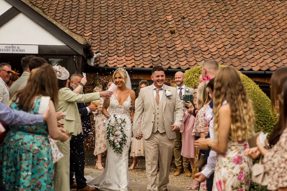 Wedding party and guests gathered outside rustic venue on sunny day.