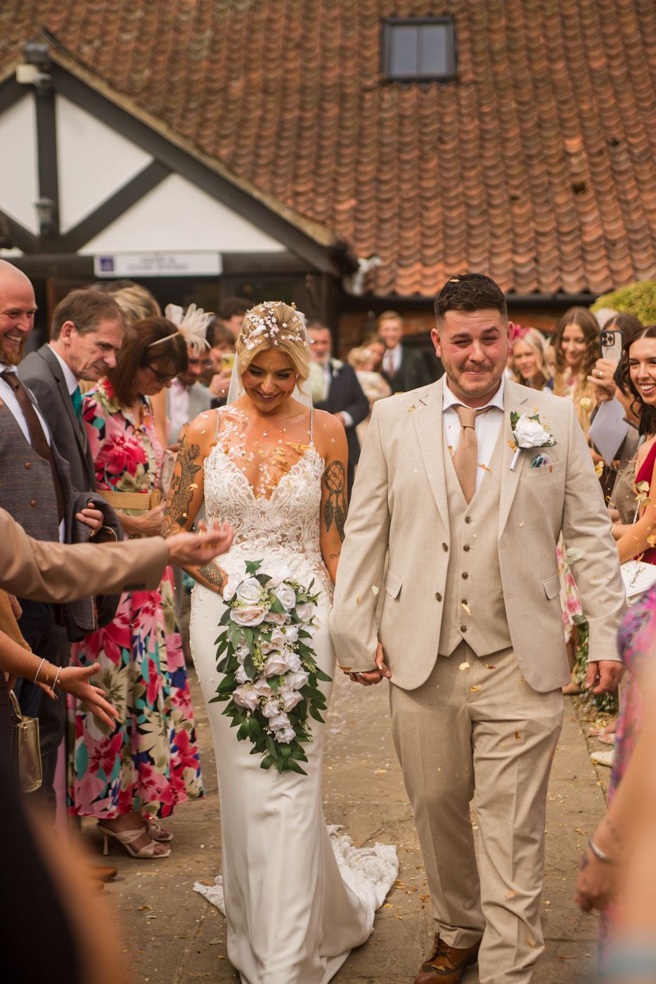Wedding party wearing beige suits and white dresses walking together at an outdoor venue with terra cotta roof tiles.