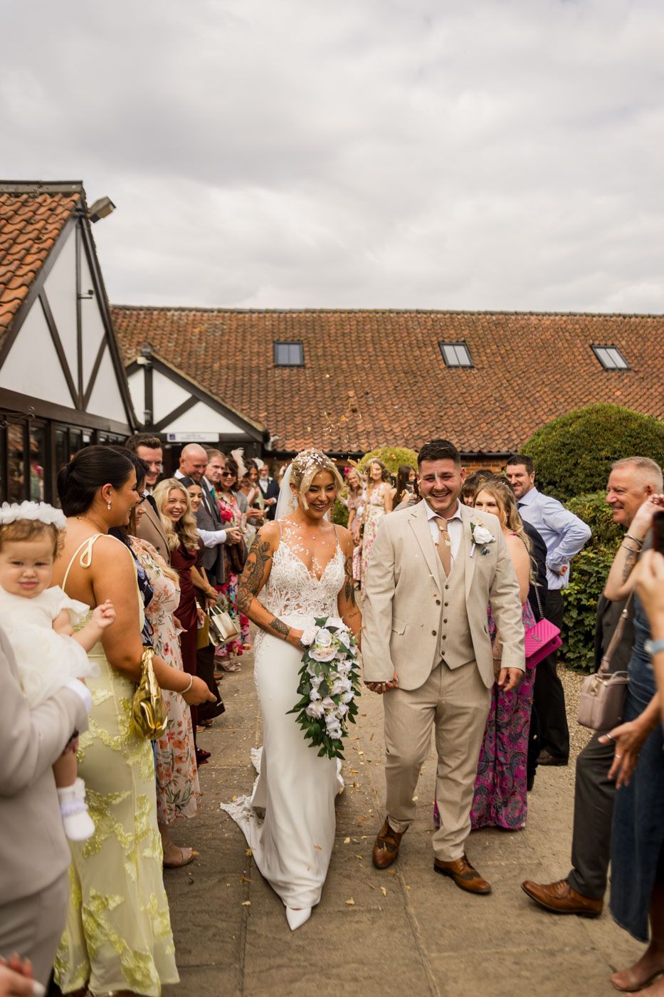 Wedding party walks through guest-lined path outside venue under cloudy skies.