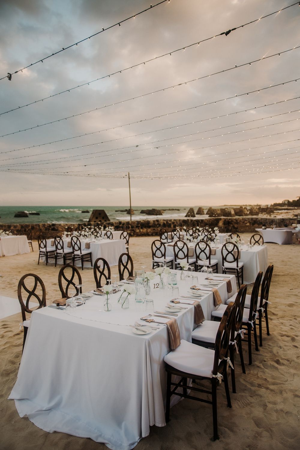 Elegant beachside wedding reception setup with white tablecloths and dark wooden chairs on sandy shore at sunset. El Dorado Royale Wedding