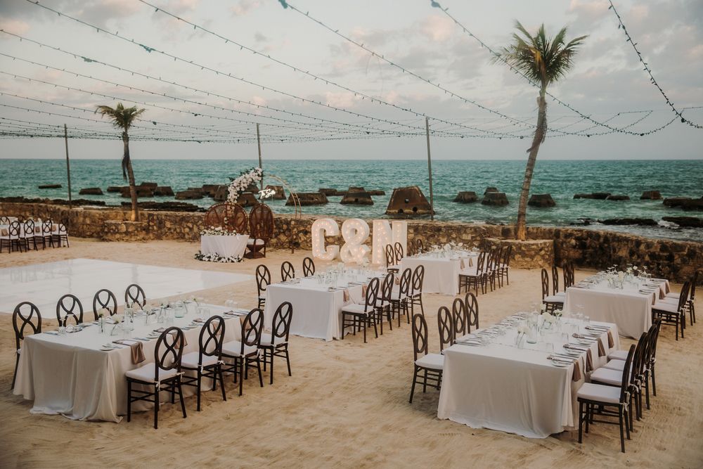 Elegant beach wedding reception setup with wooden chairs and white tables overlooking the ocean at sunset. El Dorado Royale Wedding Photo