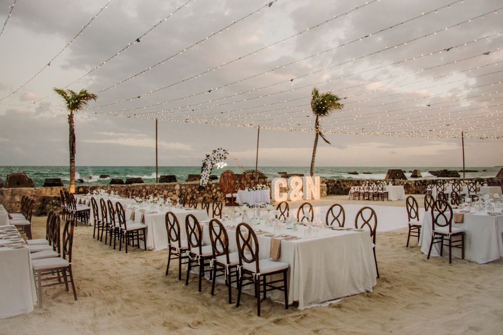 Elegant beach wedding reception setup with white linens and dark wooden chairs arranged on pristine sand at sunset. El Dorado Royale Wedding