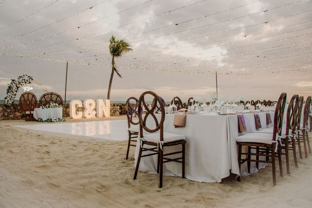 A long white dining table with wooden chairs set up on a sandy beach at sunset for an elegant outdoor reception. El Dorado Royale Wedding