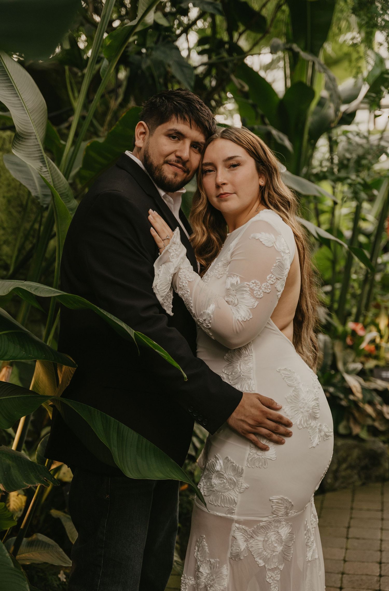 A couple poses together in a lush indoor greenhouse conservatory filled with tropical plants and palm fronds.