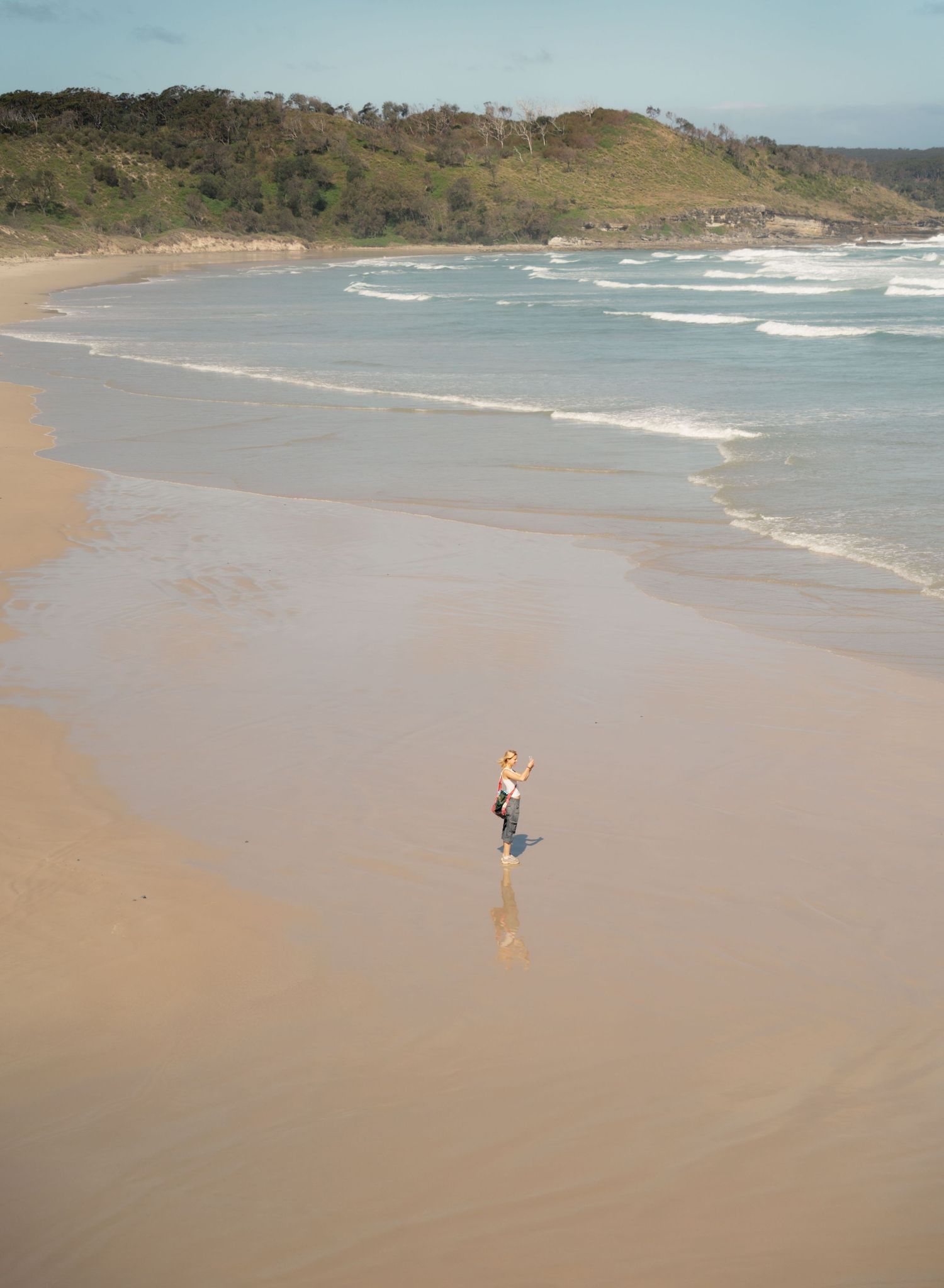 A lone figure walks along a vast empty beach with rolling hills in the background.