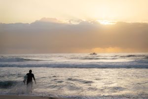 A silhouetted surfer wades through ocean waves at sunset with mountains in background.
