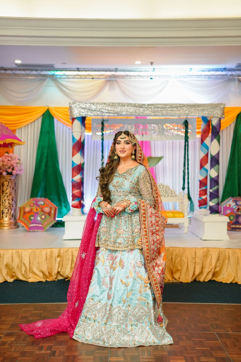 Ornate South Asian bridal lehenga in mint blue and pink with detailed embroidery at a decorated wedding venue stage.