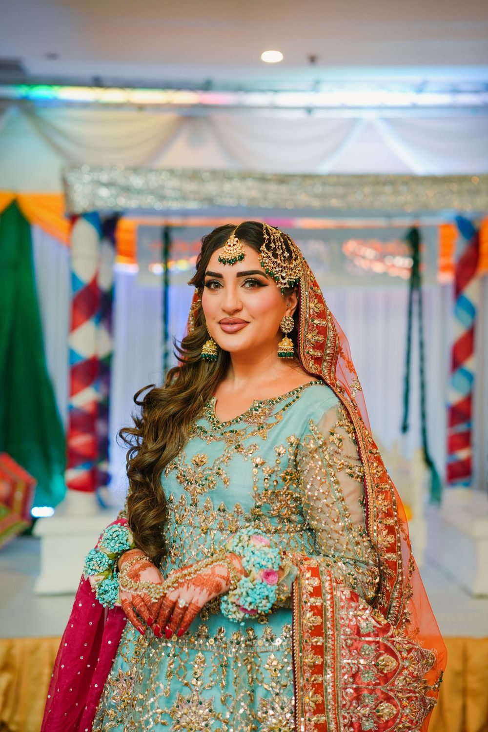 A bride in a stunning turquoise and red traditional South Asian wedding outfit with embellished jewelry and long wavy hair.