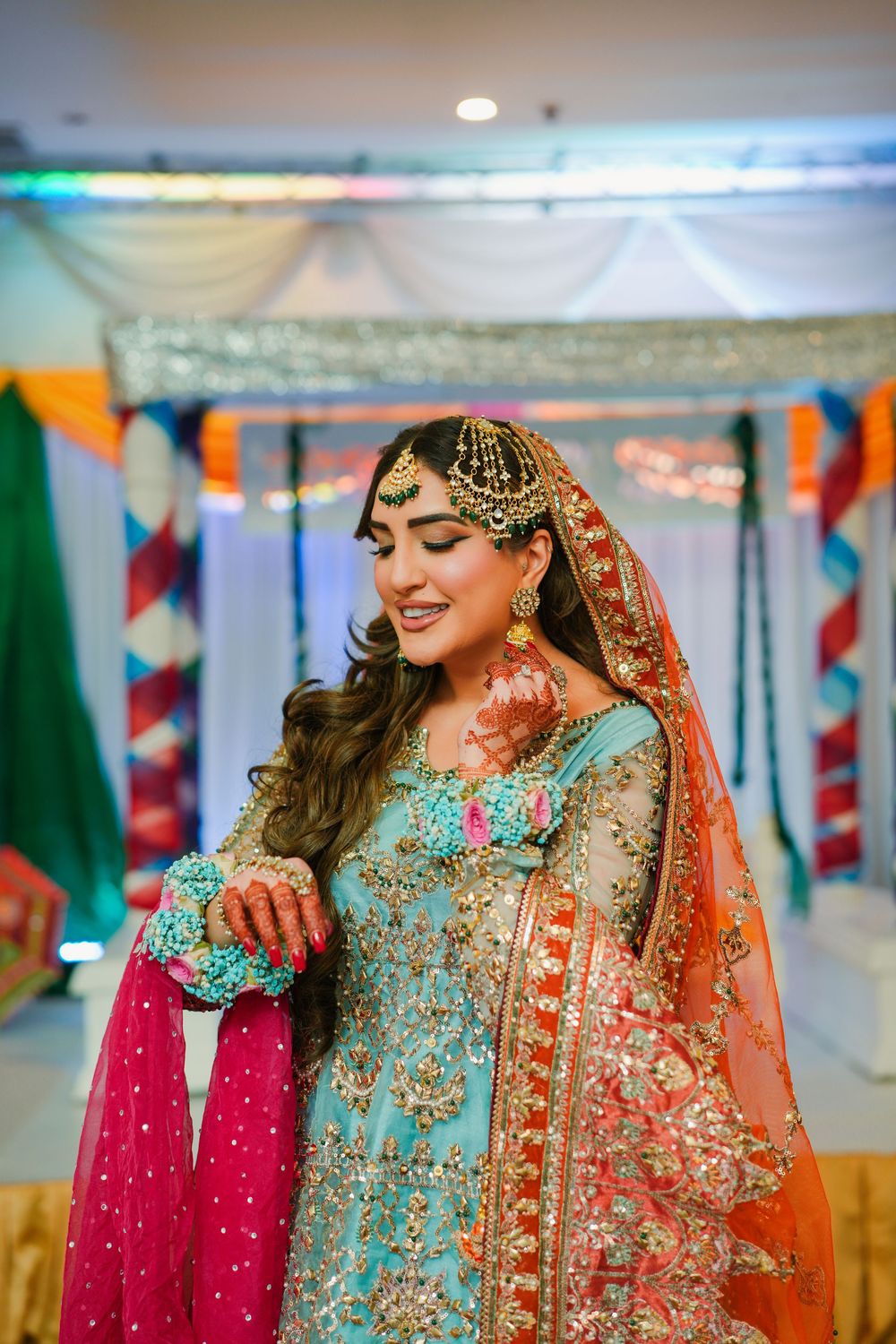 Beautiful bridal attire featuring an embroidered turquoise dress with orange dupatta and ornate gold jewelry at a festive event.