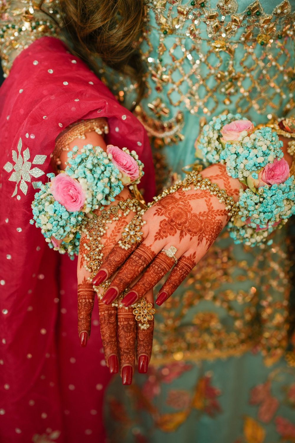 Hands adorned with intricate henna designs hold turquoise and pink flowers against a red and gold traditional outfit.