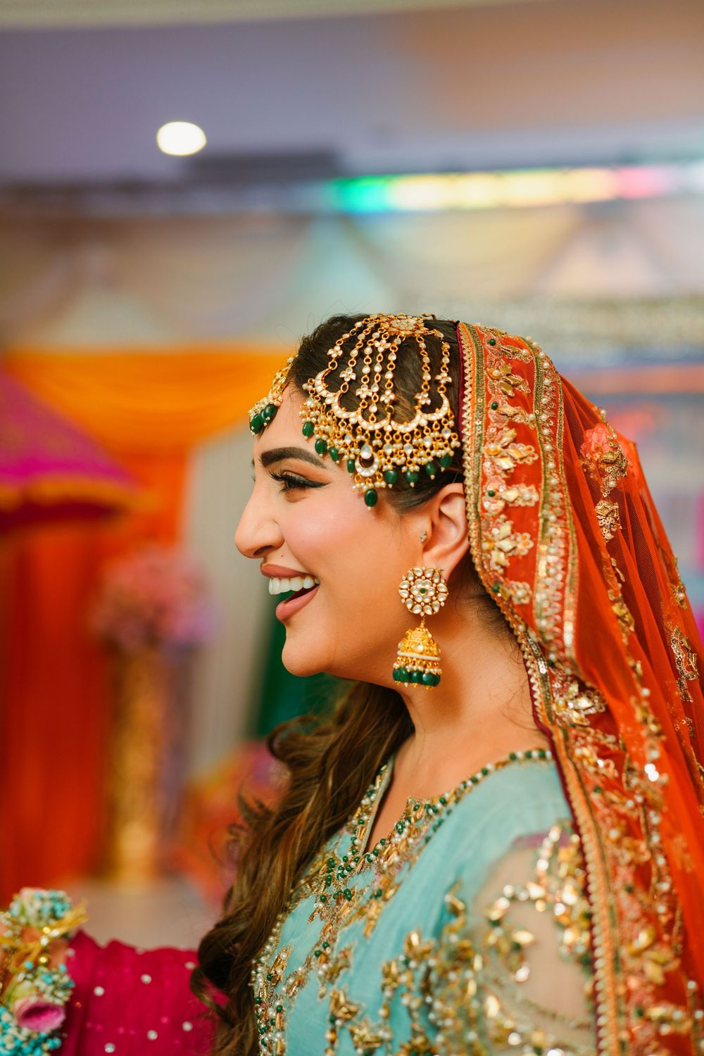 Traditional South Asian bride wearing ornate jewelry and orange dupatta smiles during wedding celebration.