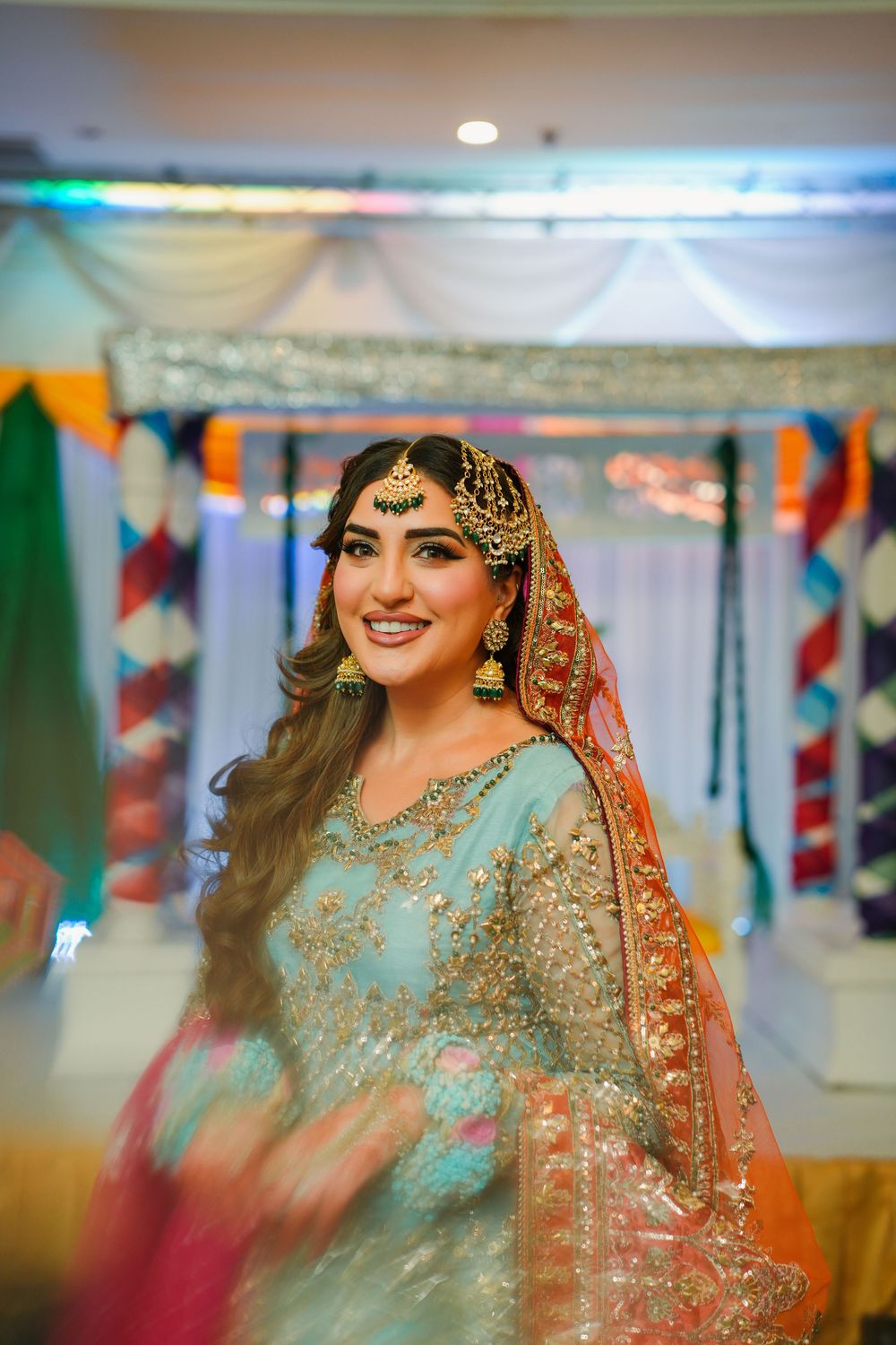 Traditional Indian bride wearing a turquoise blue lehenga and ornate gold jewelry in a decorated wedding venue.