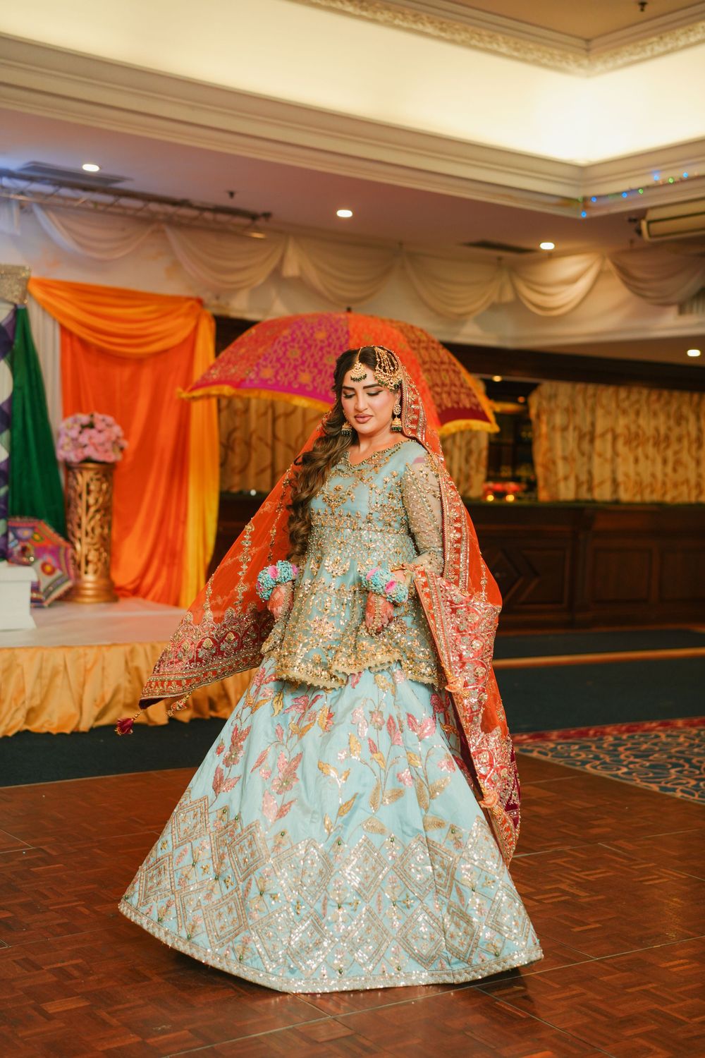 Bride in light blue and coral traditional lehenga poses with decorative umbrella at wedding venue.