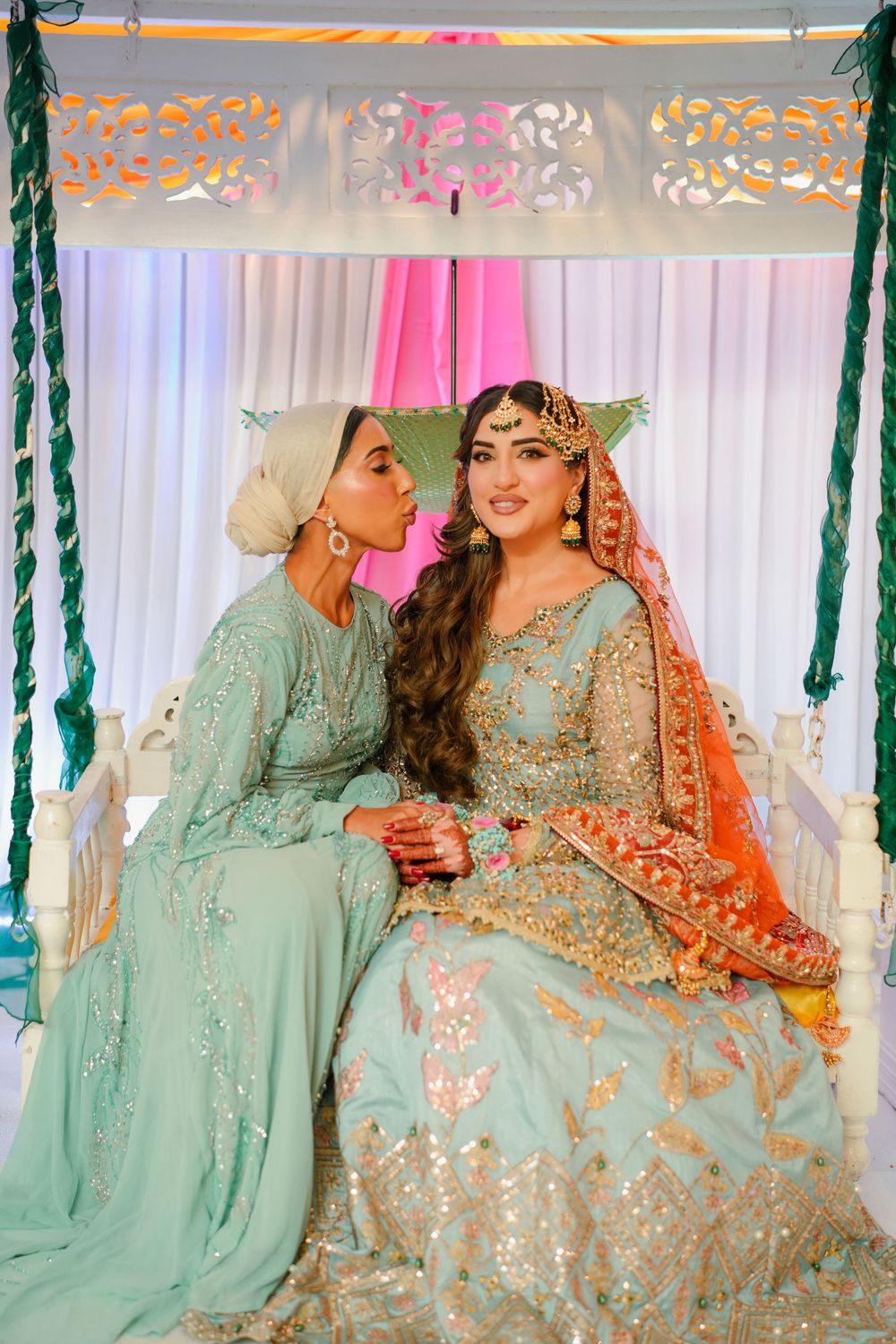 Two women in traditional South Asian outfits share a moment under decorative canopy with mint and coral colors.