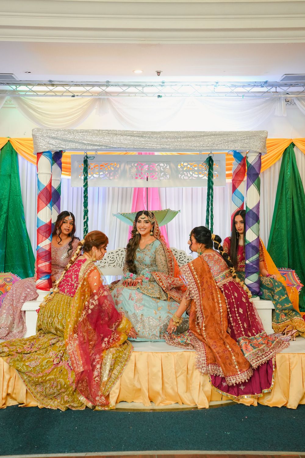 A group sits arranged in colorful lehengas and traditional Indian attire during a festive celebration or wedding event.