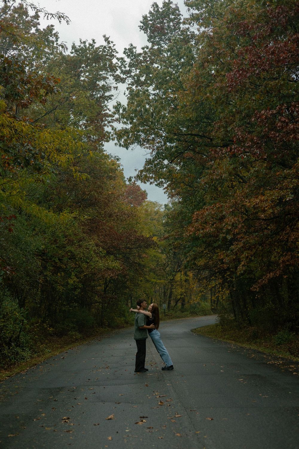 A wide cinematic photo of a couple embracing in the autumn leaves on a trail in Brighton, Michigan.