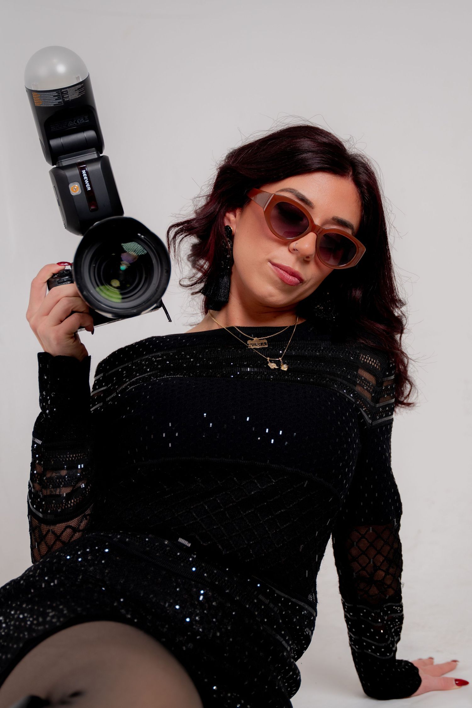 Professional photographer with dark curly hair wearing a black t-shirt holds a camera while smiling against a brick wall.