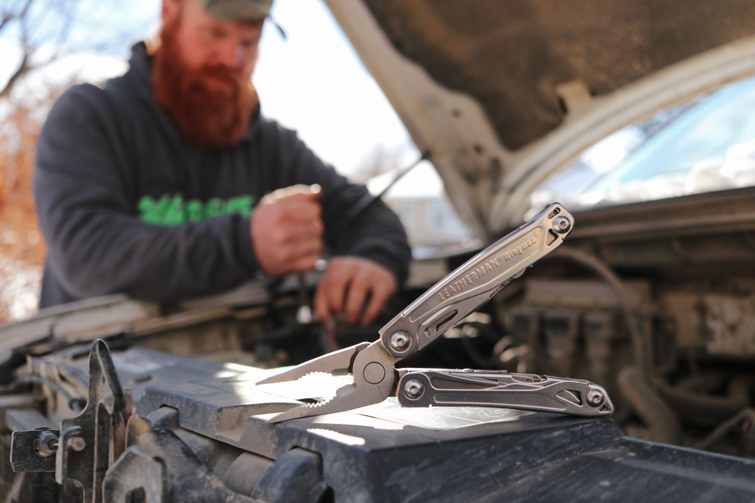 Multi-tool pliers resting on car engine while mechanic works in background.