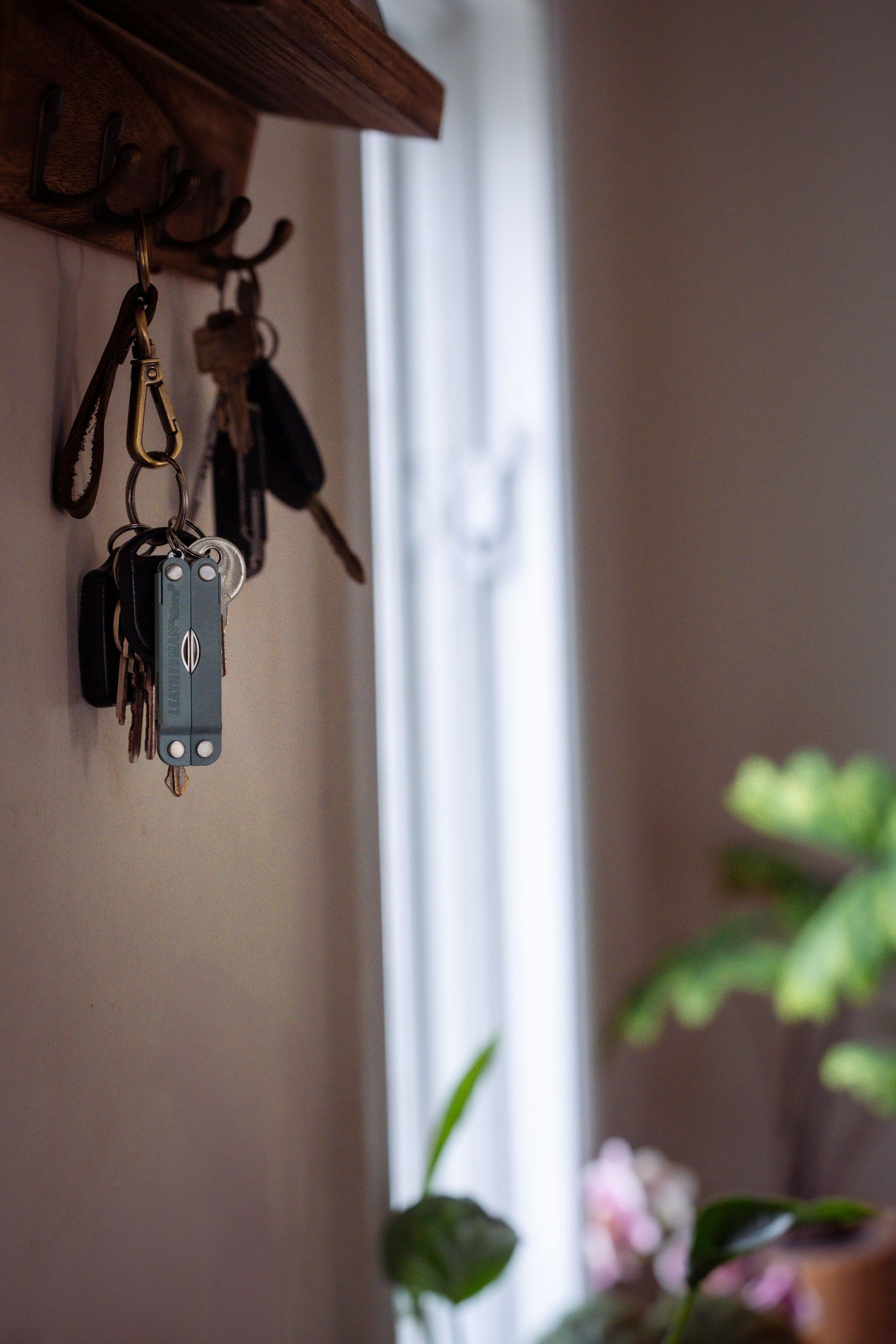 Multi-tool keychain hanging from wooden wall hook near window with plants.