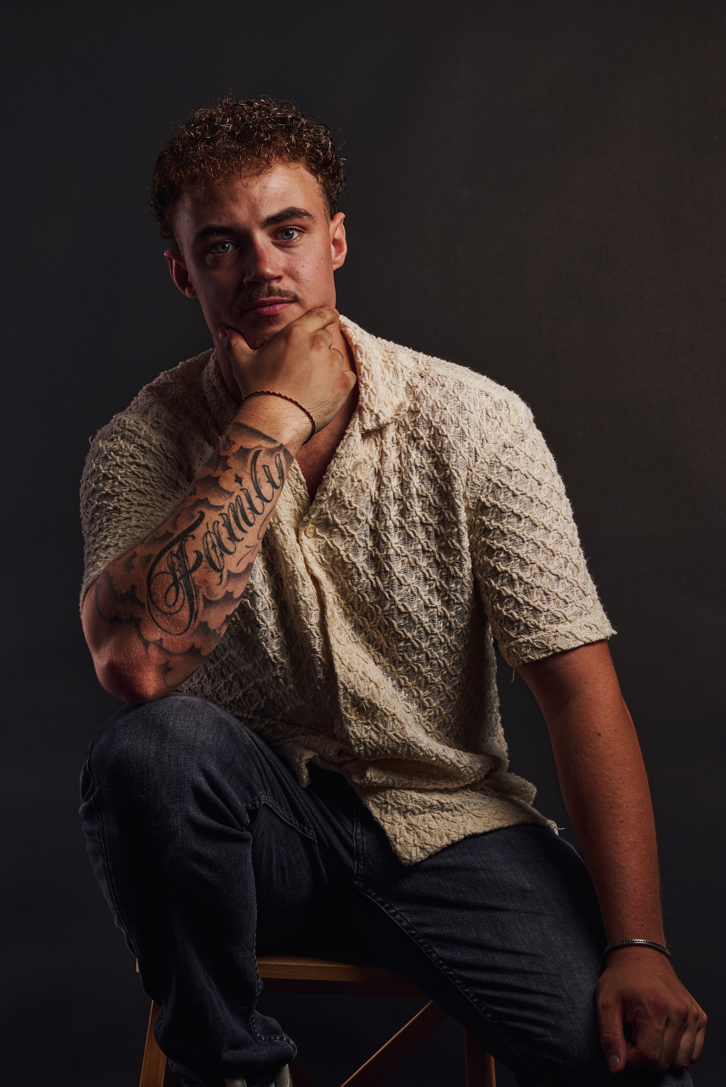 Jonge man met krullend haar en voorarm tattoo, zittend op een stoel in een nadenkende houding op een zwarte achtergrond.