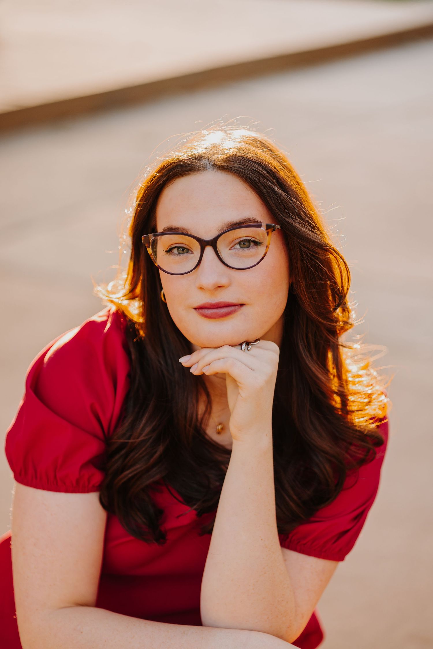 A woman in a red dress and glasses poses thoughtfully against a neutral background in warm lighting.
