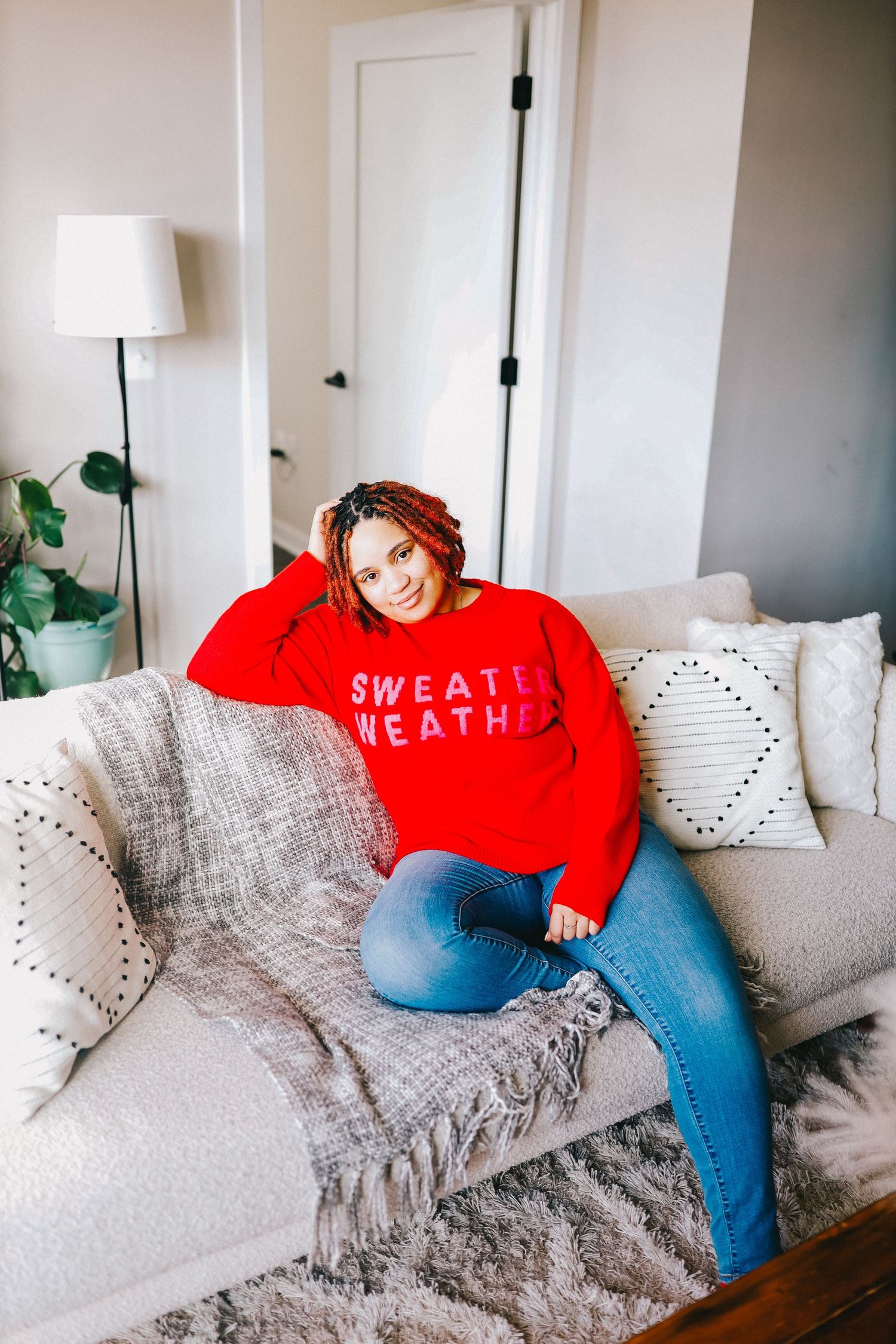 Casual lifestyle photo in cozy living room with red sweater and jeans on gray couch with decorative throw pillows.