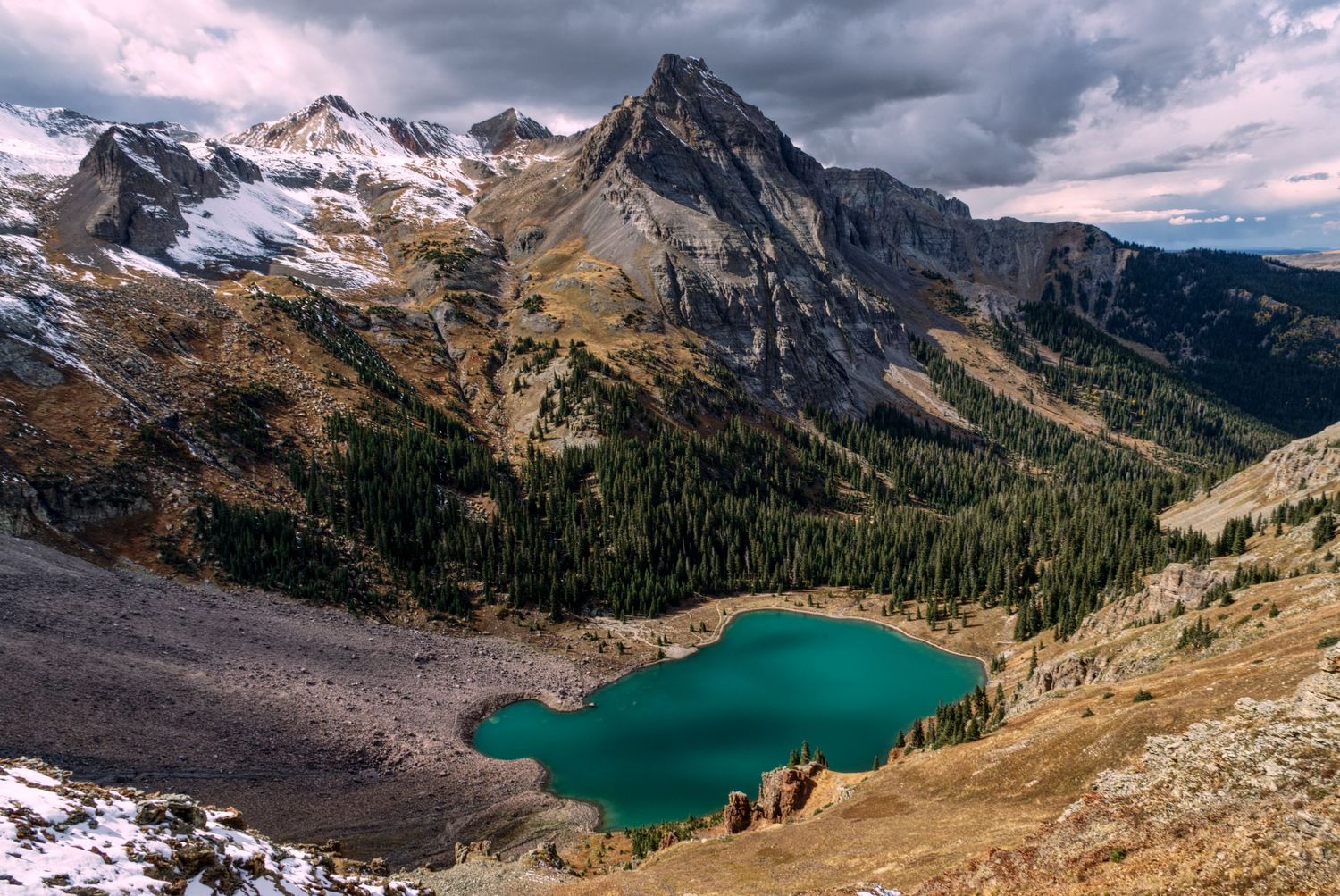 Towering  snowy mountain peaks surrounded by a dramatic sky over a pretty lake below.