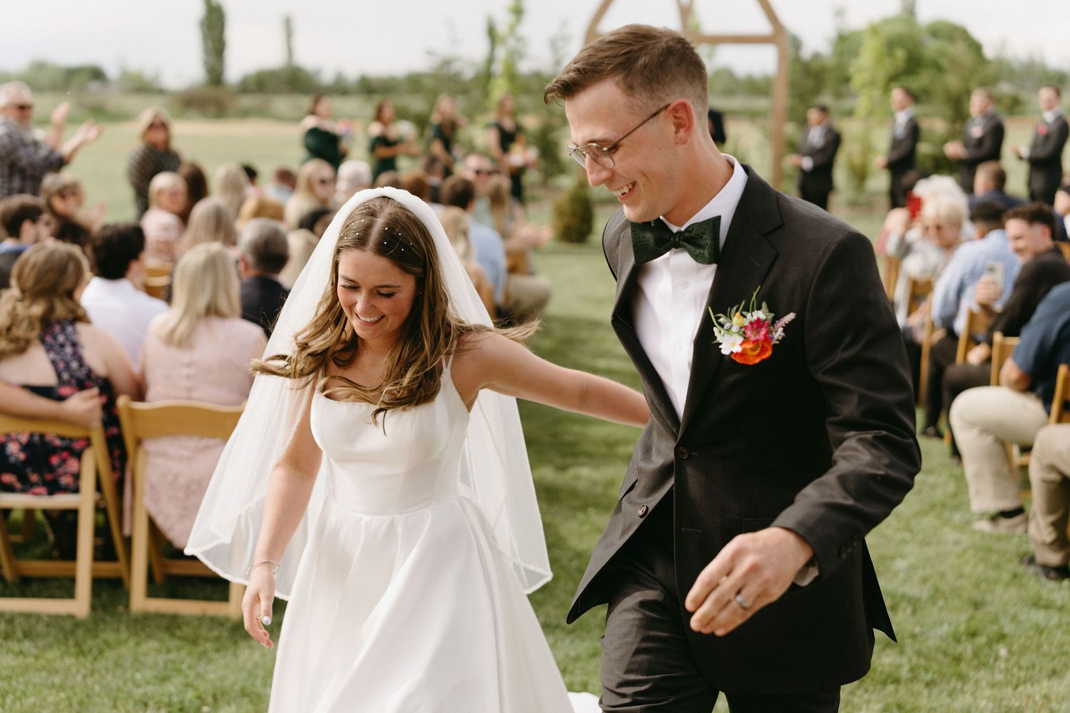 Couple walks together during outdoor wedding ceremony with guests seated on wooden chairs in garden setting.