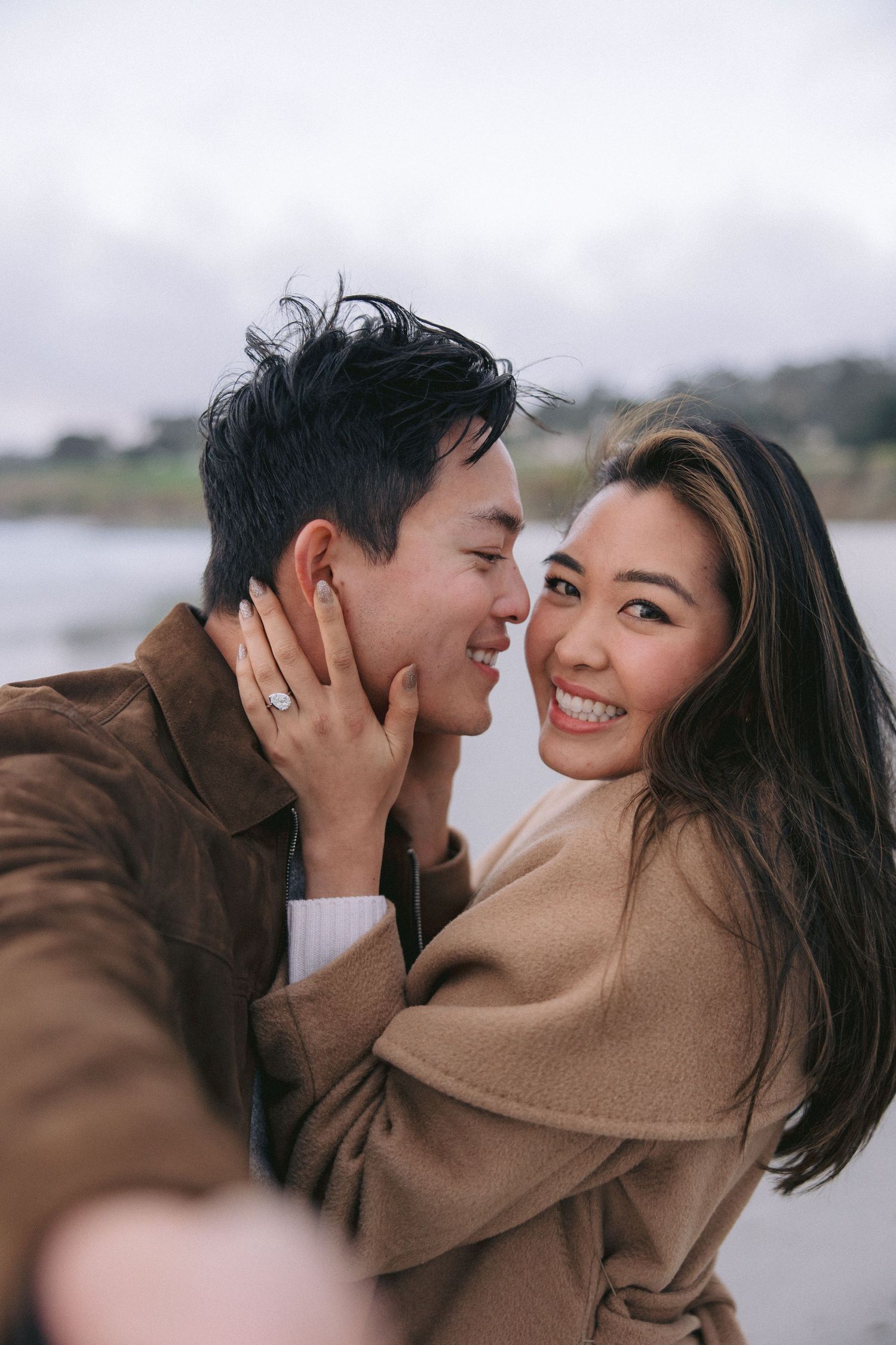A romantic couple in brown coats embrace and smile together by a waterfront on an overcast day.