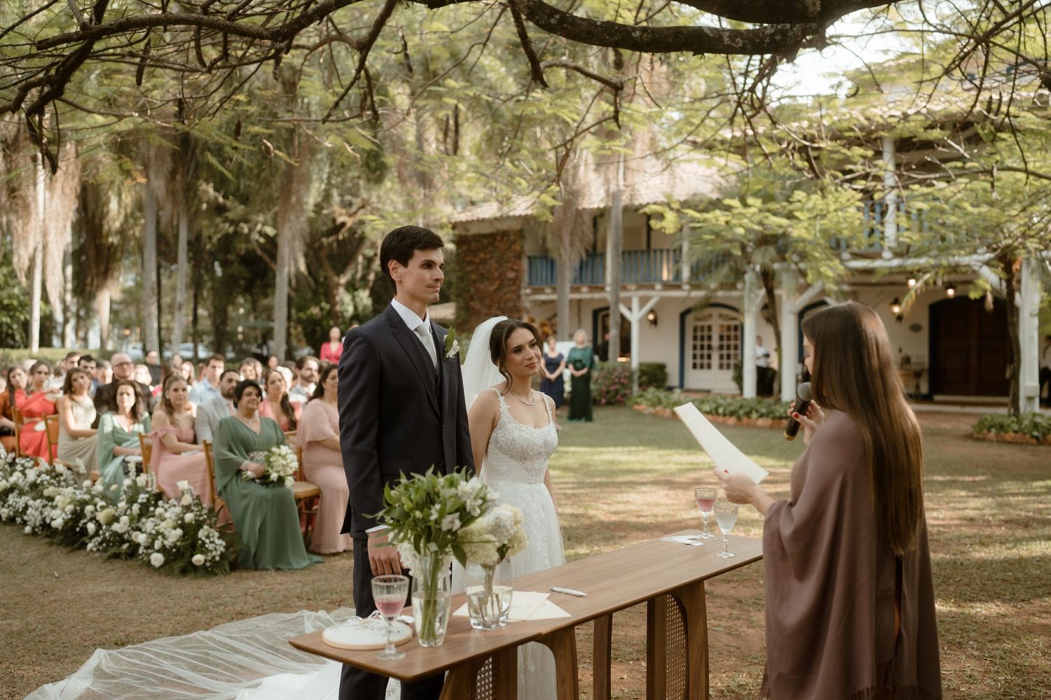 Panoramic view of outdoor wedding ceremony setup with guests seated under tree canopy.