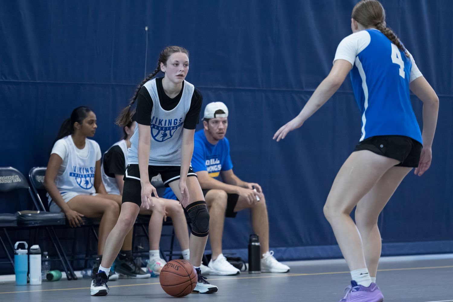 A basketball player dribbles while teammates watch from the sideline during an indoor practice session.