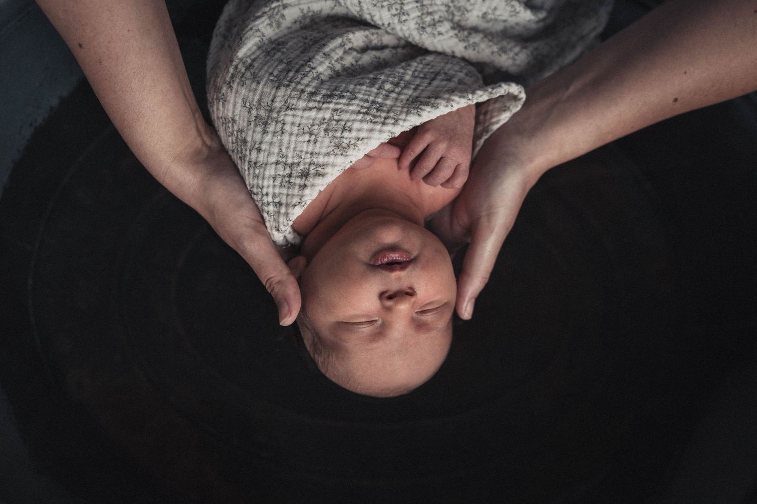 Vue en plongée d’un bébé emmailloté dans un lange gris, entouré de mains délicates, plongé dans son thérapeutique bain bébé.