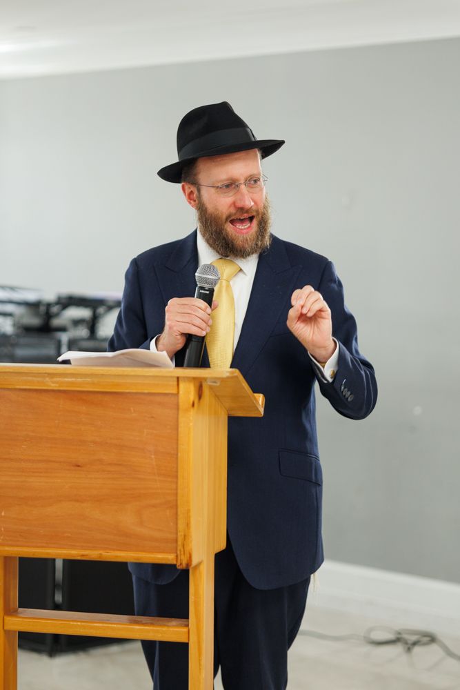 A speaker in a navy suit and yellow tie gestures animatedly while standing at a wooden podium.