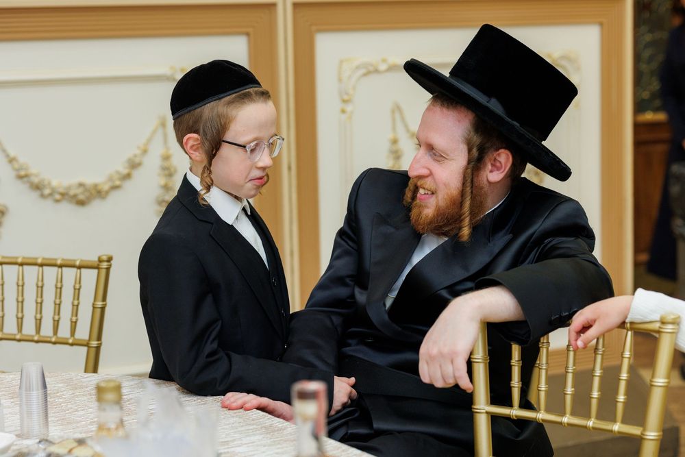 Two Orthodox Jewish men having an animated conversation while seated at a table with gold chairs.