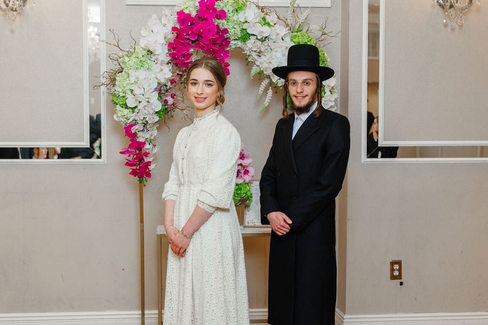 Young Orthodox Jewish couple in formal attire posing before a floral arch with pink and white flowers.