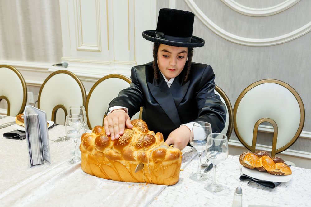 Young Orthodox Jewish man in black coat and top hat tearing challah bread at a festive Shabbat table.