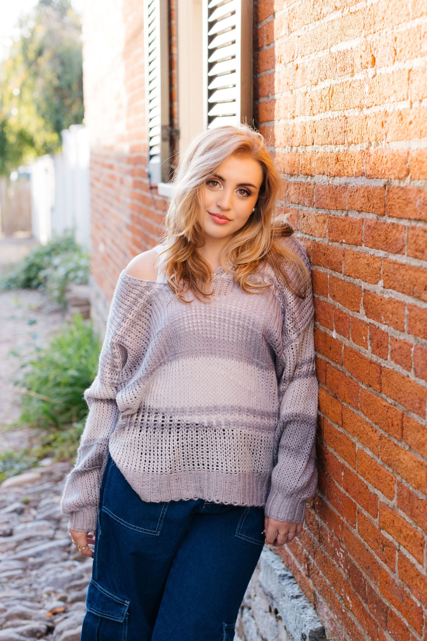 Young woman in grey knit sweater leans against brick wall on cobblestone path outdoors.