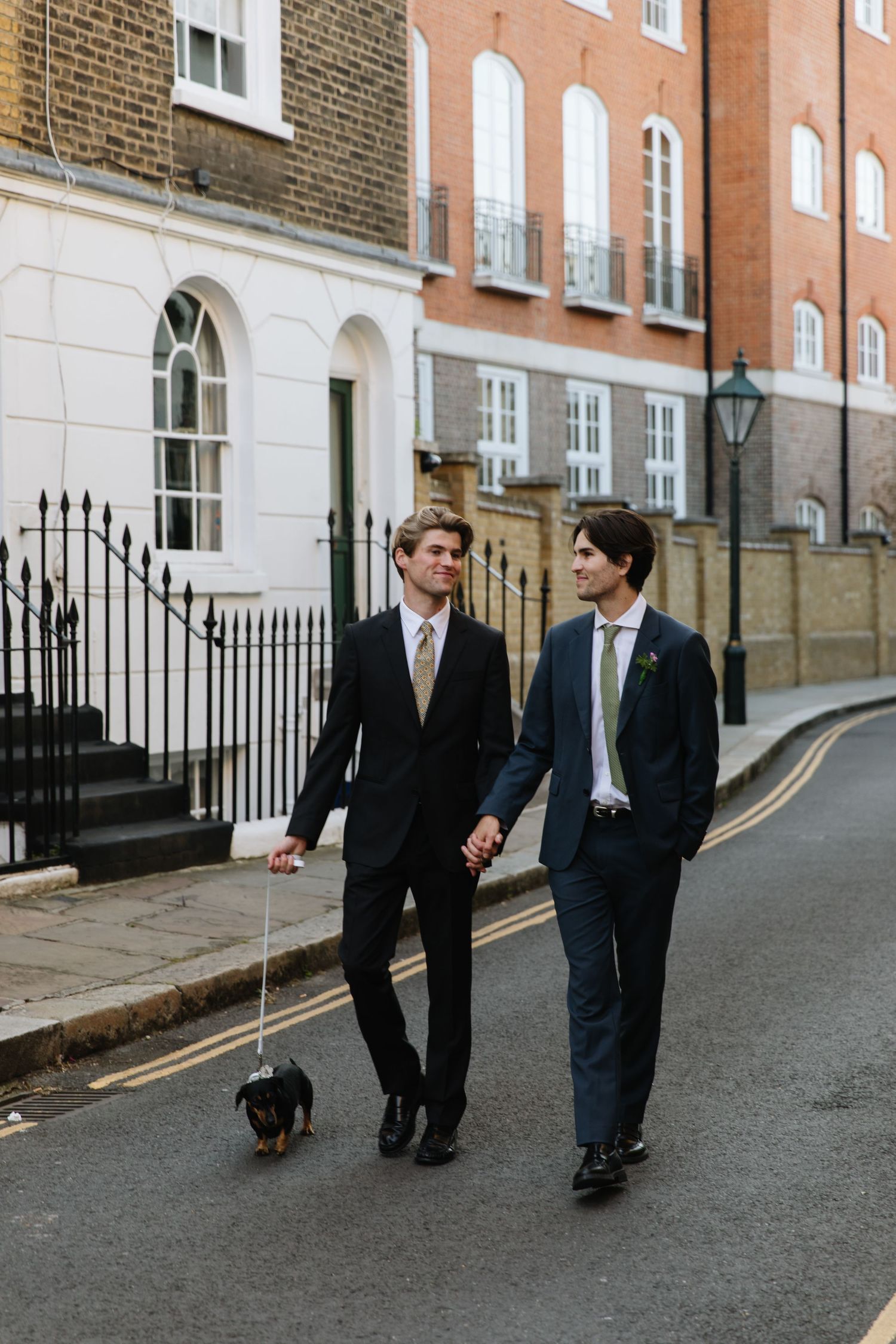 Two people in suits walk a small dog down a classic London street.